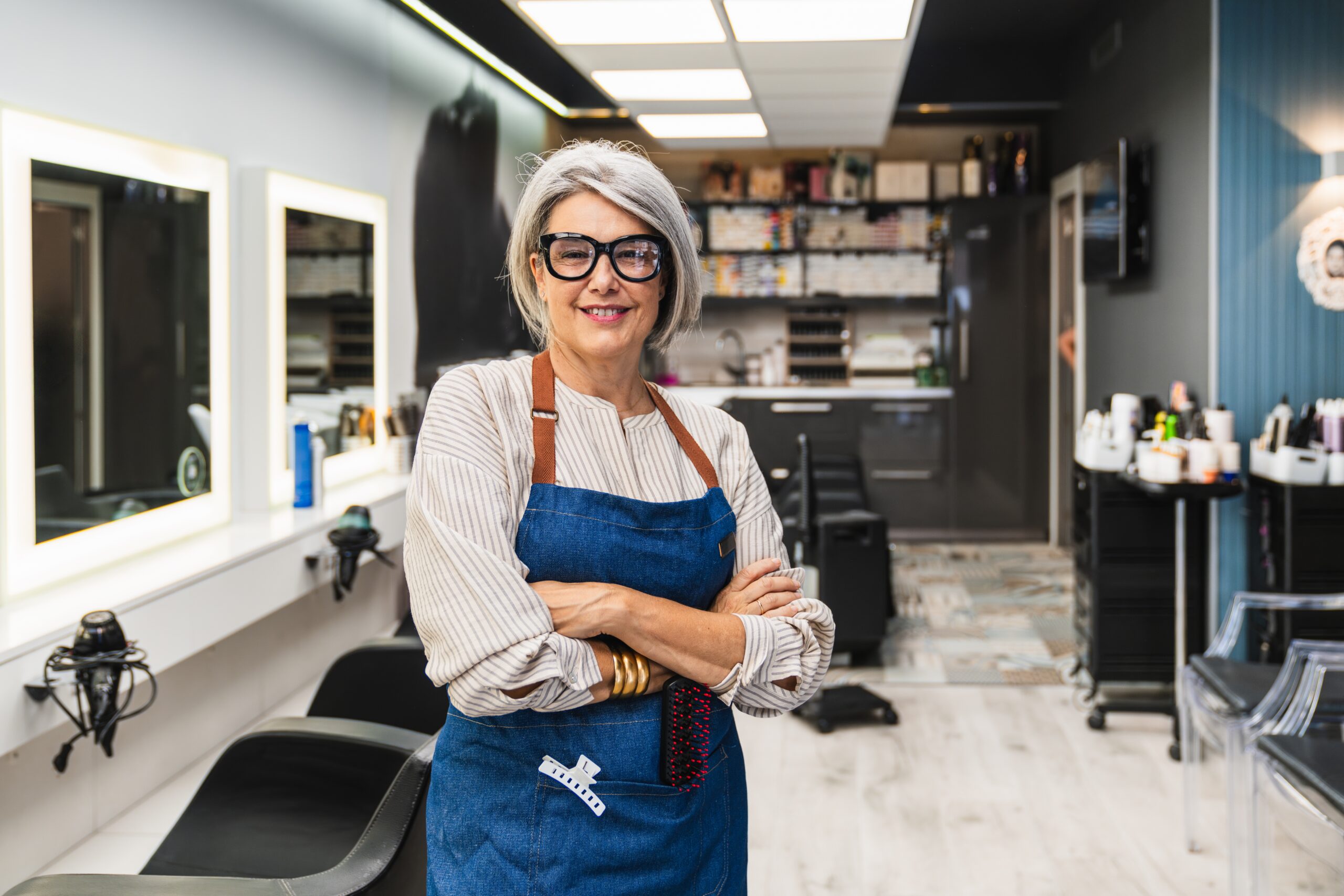 Senior woman entrepreneur standing with arms crossed, smiling inside her modern hair salon. Successful small business owner portrait.